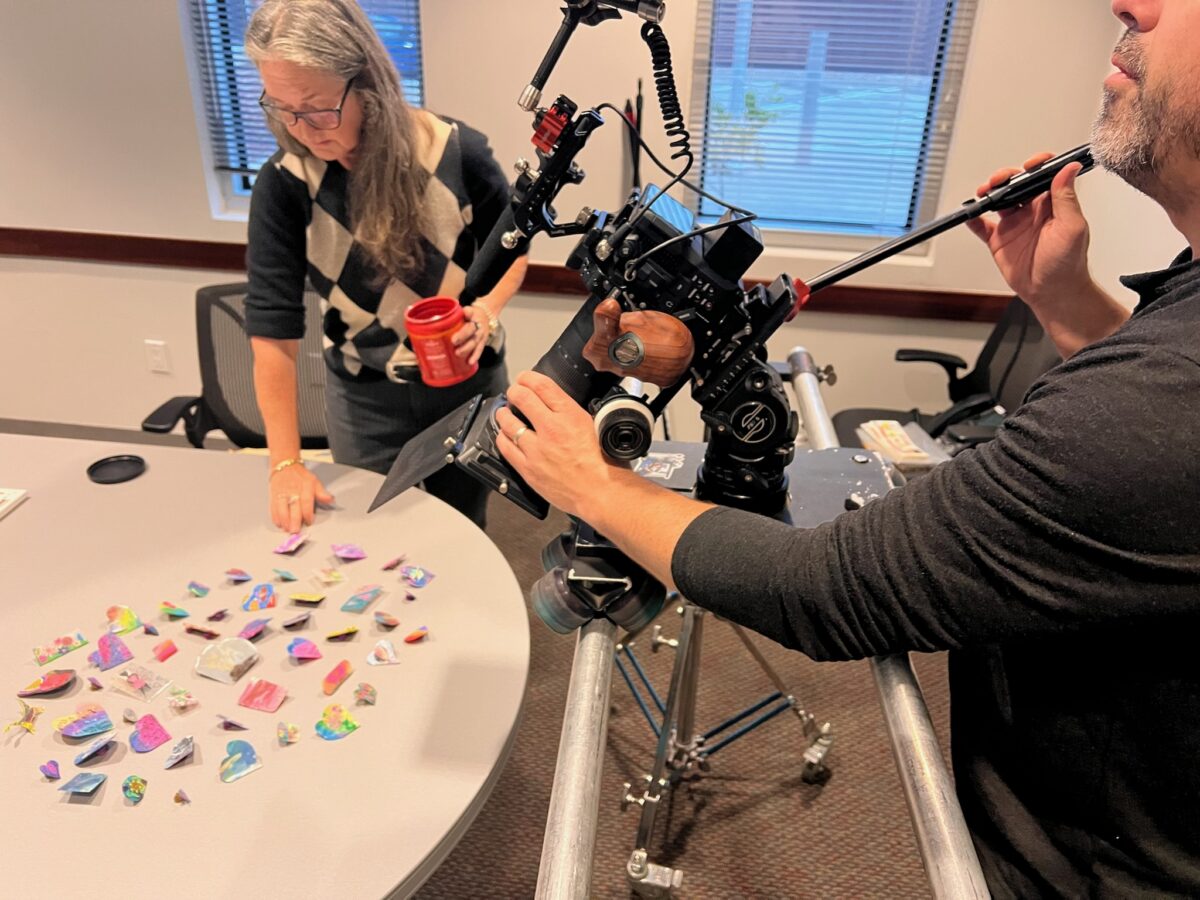Camera operator films a closeup of small items on a table while a woman helps arrange them, behind the scenes of Cut To Create's project for Rice University's Kinder Institute for Urban Research