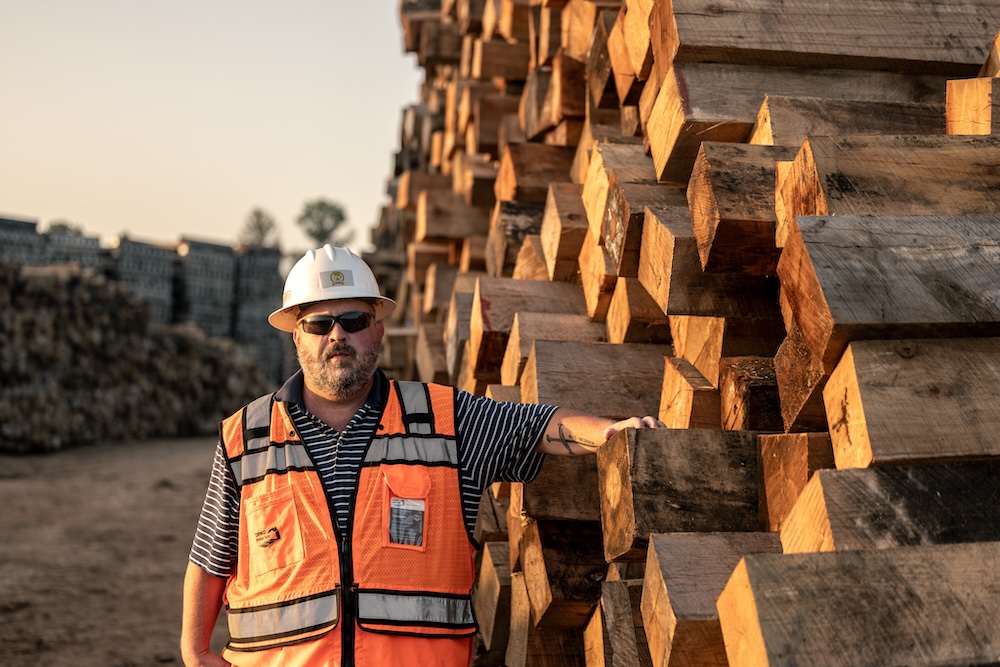 A man with safety gear and a forearm tattoo leans on a wall of unstacked timber.