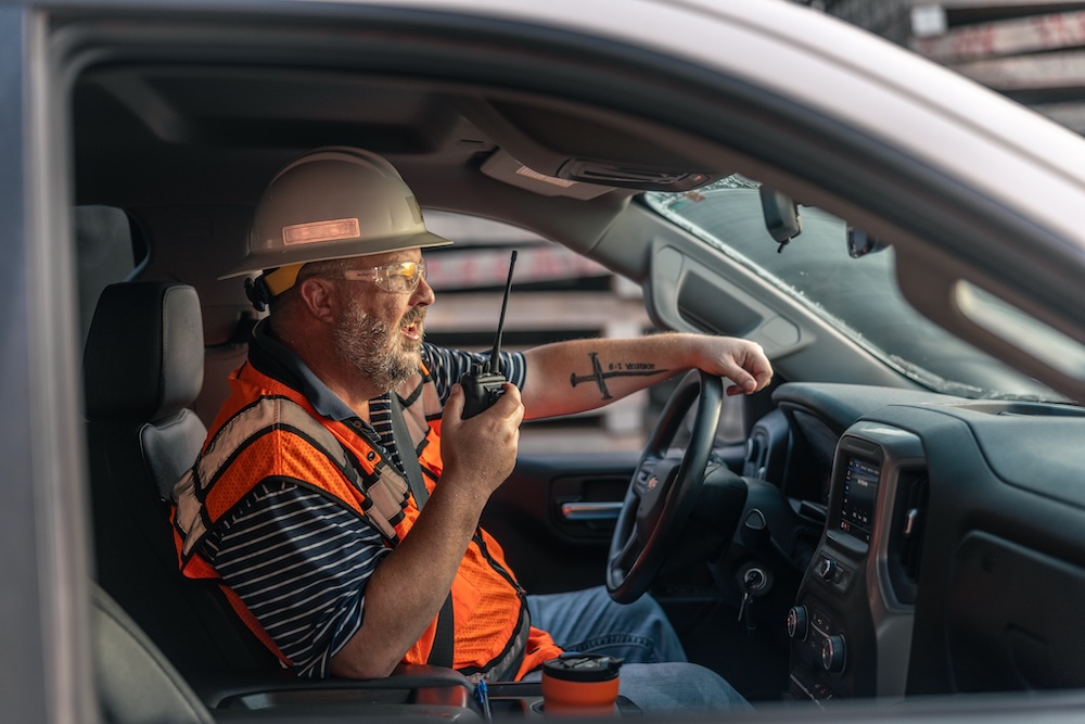 A man with a cross tattoo on his forearm wearing an orange safety vest, safety glasses and hard hat sits at the wheel of a vehicle talking on a handheld radio.