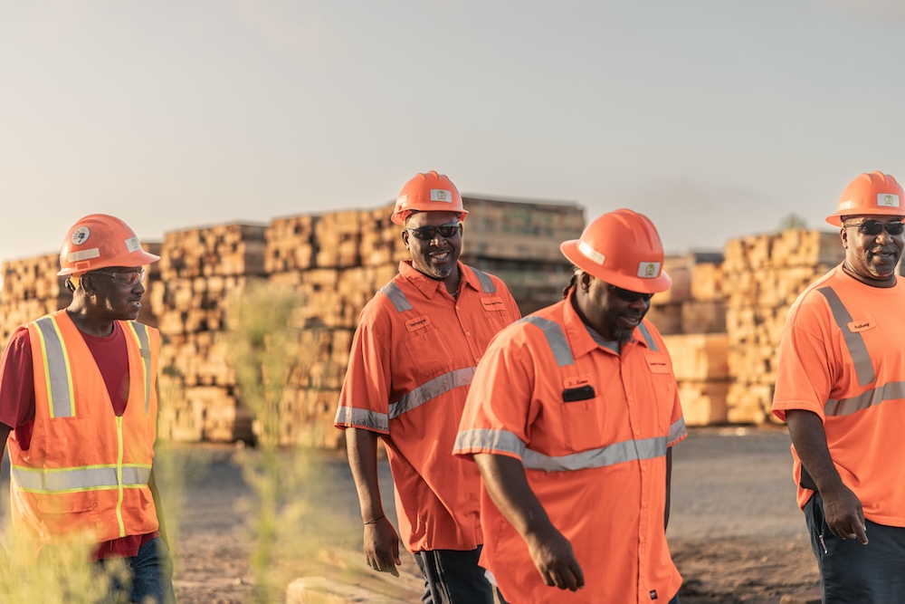 Four men in orange safety shirts and hard hats walk and laugh in front of tall stacks of wood.