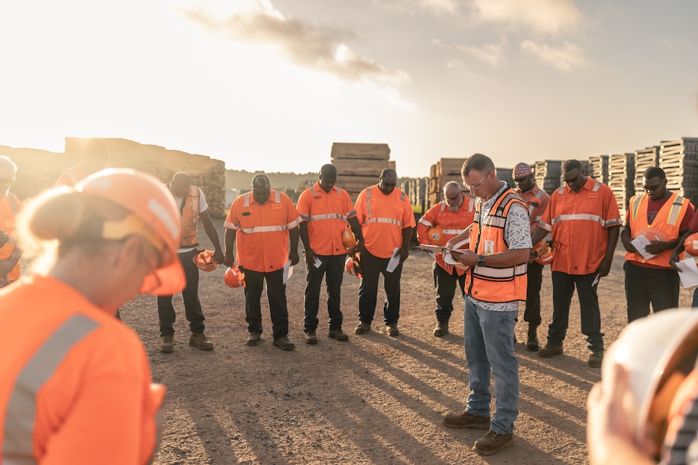 A group of workers in safety gear stand in a circle while a man in the middle reads from a clipboard.