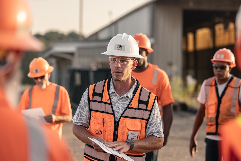 A man in safety gear gestures to his clipboard while other workers look on.