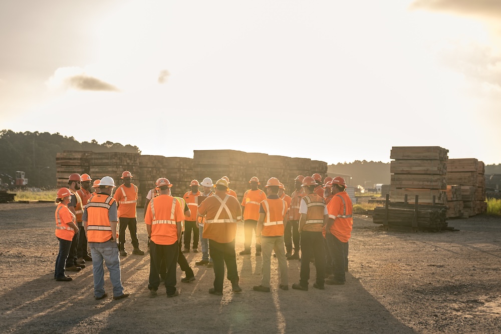A group of workers in safety gear meet in a gravel lot with tall stacks of timber in the background.