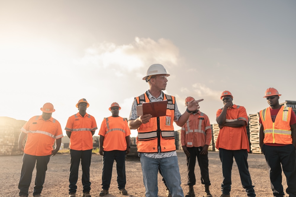 A line of workers in safety gear stand behind a worker also in safety gear holding a clipboard and gesturing to his left.