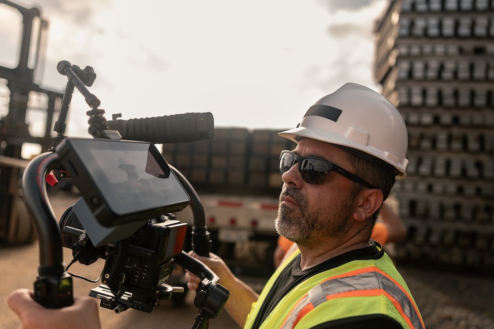 A camera operator in hard hat and glasses views his monitor as he films another worker off-camera.