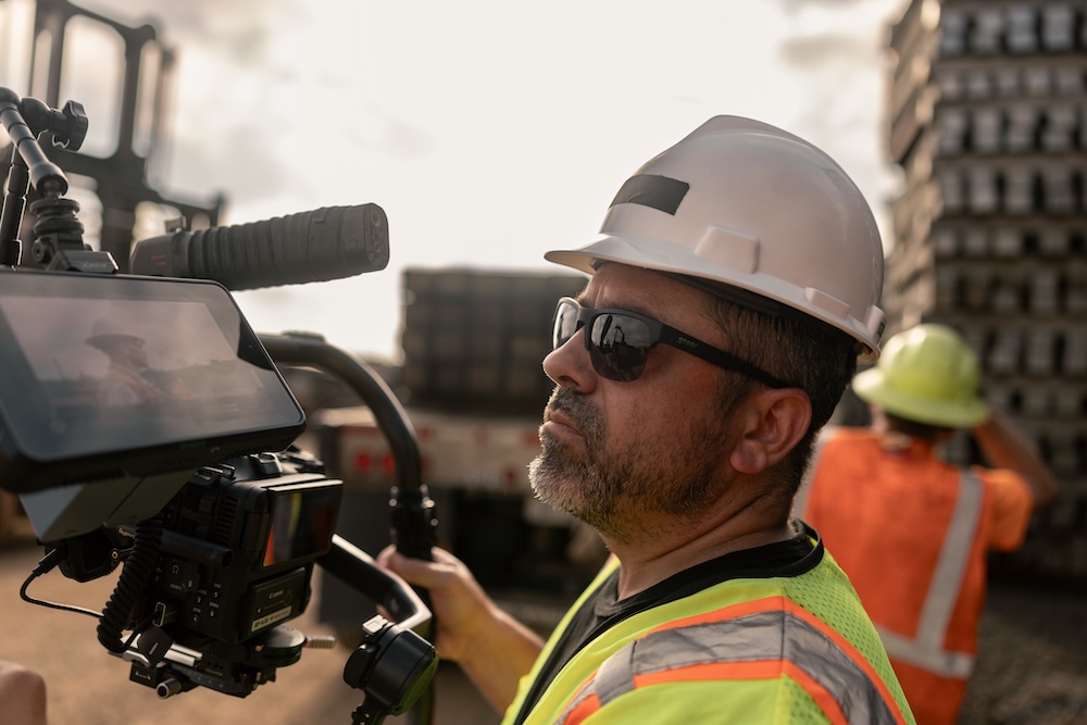 A camera operator in a hard hat and sunglasses is looking at his camera monitor while a worker loads timber in the background.