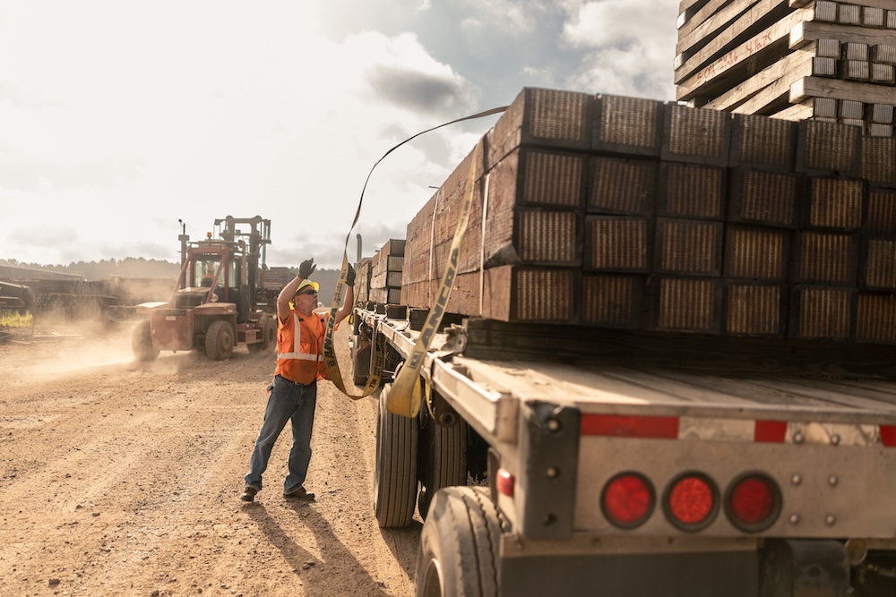 A worker in safety gear is throwing a strap over a stack of timber on a semi truck bed.