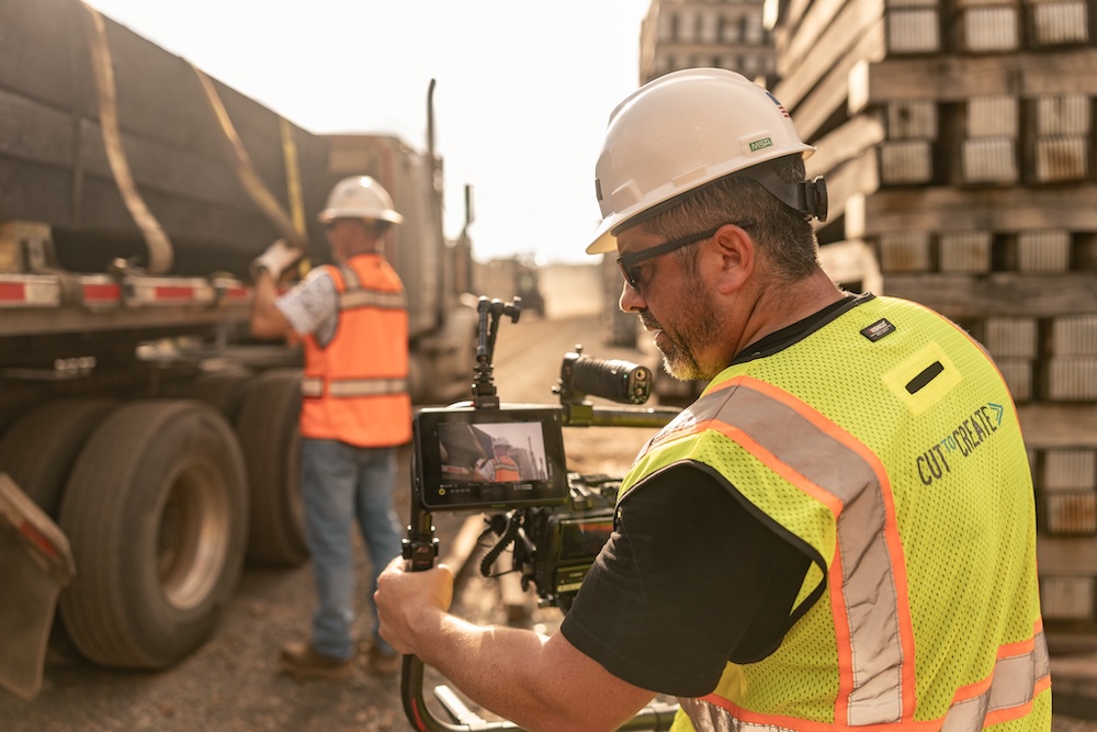In the foreground a camera operator in a safety vest and hard hat is filming a worker in the distance who is loading wood onto a semi.