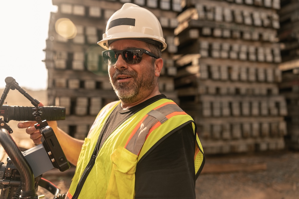 A super hip dude in sunglasses and hard hat gives a jaunty smirk as he holds his camera in front of stacks of wood.