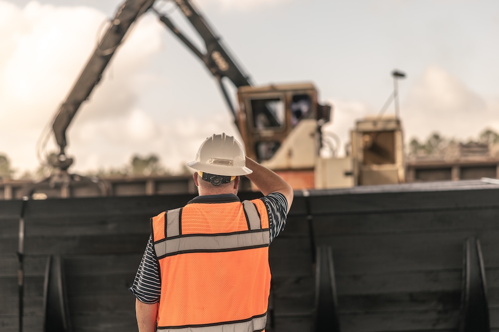 A worker in safety gear looks away from the camera toward a crane in the distance.