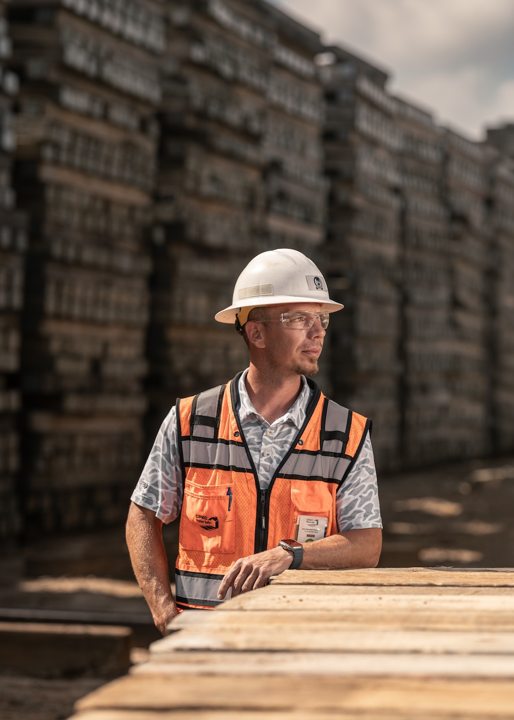 A worker in safety gear leans on a short stack of timber and looks off into the distance.