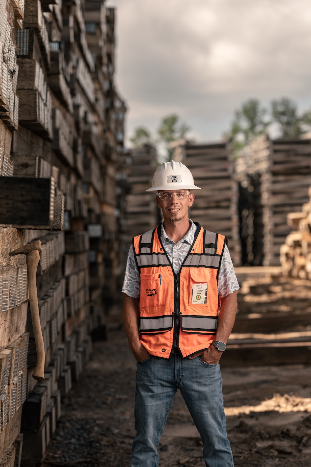 A man in a safety vest and hard hat stands with his hands in his pockets, smiling at the camera, next to a tall wall of timber with an ax stuck in the side.