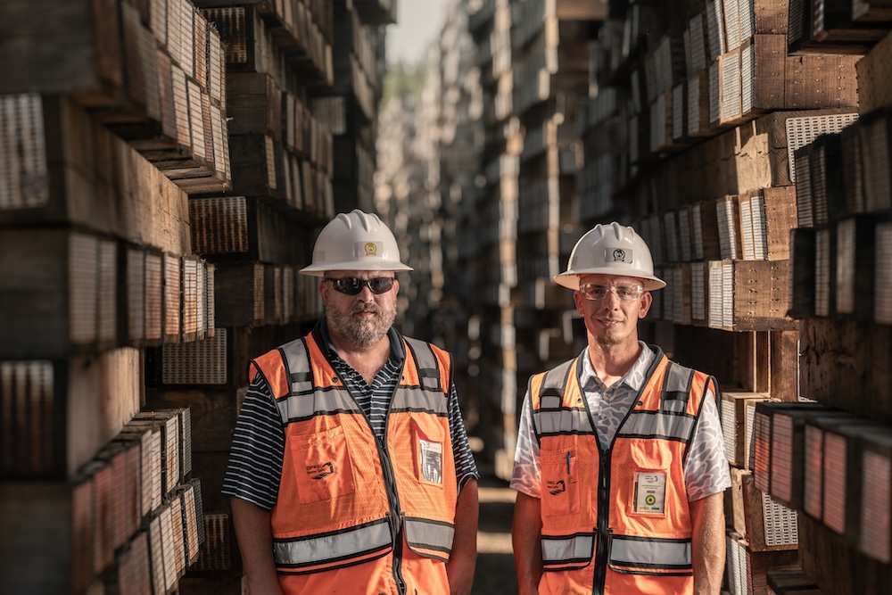 Two workers in safety vests and hard hats smile at the camera as they stand between towering walls of stacked timber.