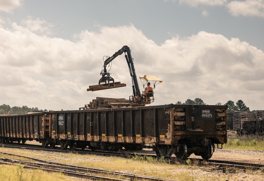 A worker uses a crane to lower pieces of timber onto a railcar.