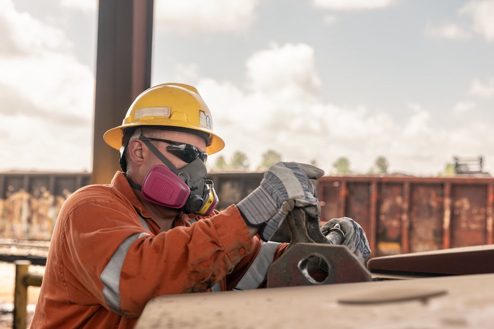 A worker in a hard hat, respirator and work gloves handles equipment in an industrial location.