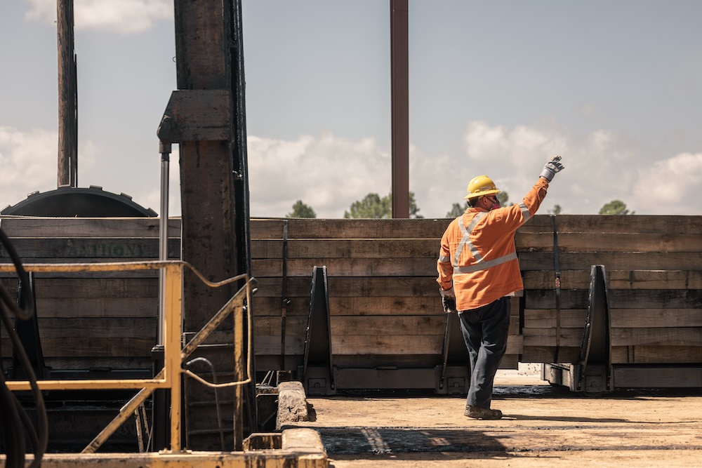 A worker in safety gear has his hand raised while large stacks of timber are being loaded for transport.