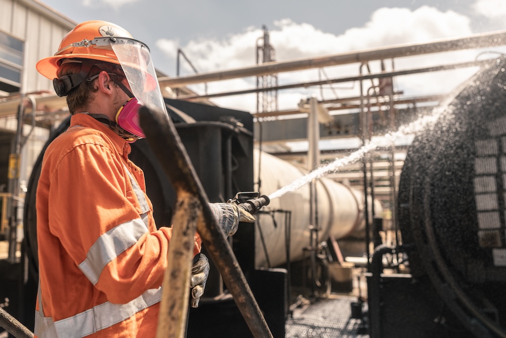 A worker in orange safety gear sprays a hose in an industrial setting.