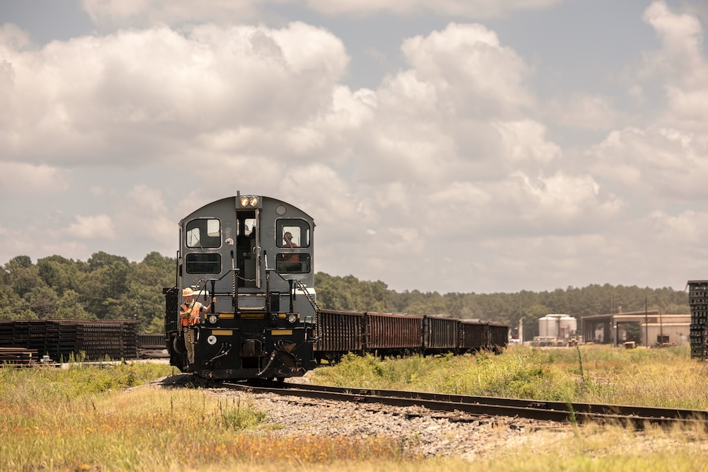 The front of a train approaches, carrying cars full of timber, with a sky full of fluffy clouds.