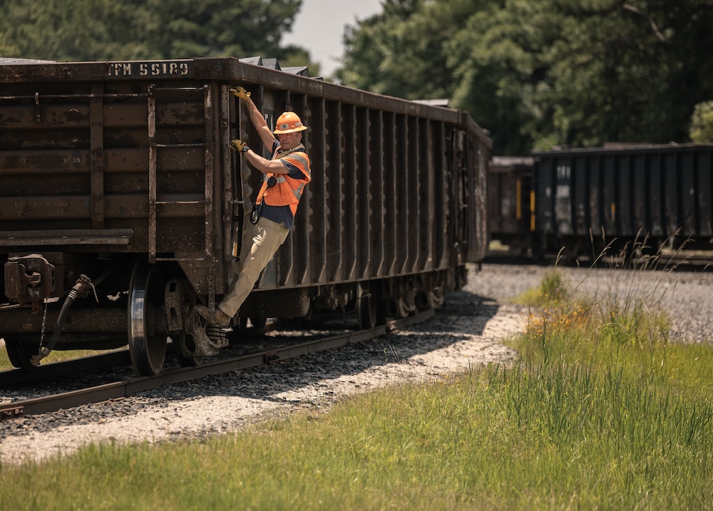 A man in an orange safety vest and orange hard hat hangs off the side of a rail car in a grassy area.