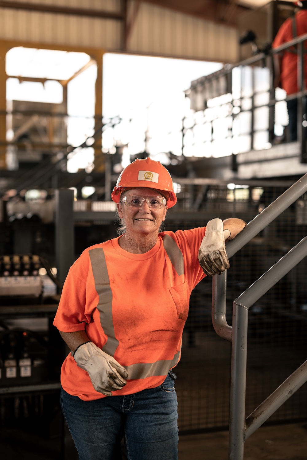A woman in an orange safety shirt, gloves, safety glasses, and orange hard hat leans on a metal rail inside of an industrial warehouse.