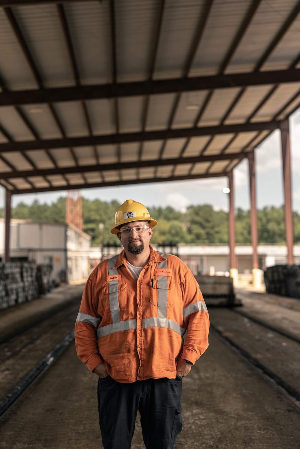 A worker in an orange safety vest, safety glasses and hard hat stands with hands in pockets smiling at the camera in an open-walled warehouse storing small stacks of timber.