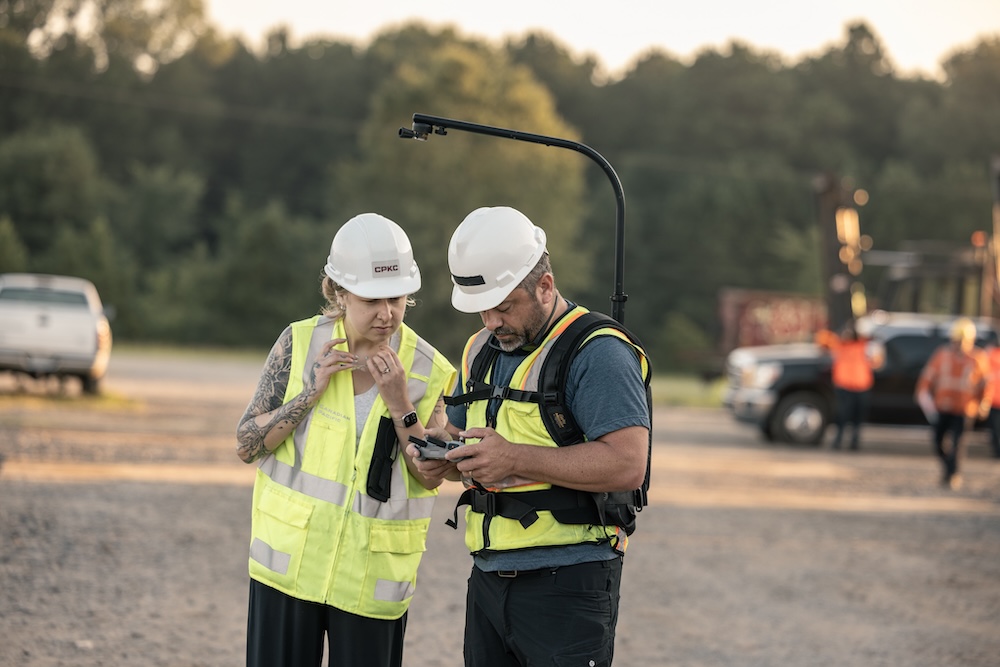 Two people in safety gear are reviewing footage in the foreground of a parking lot.