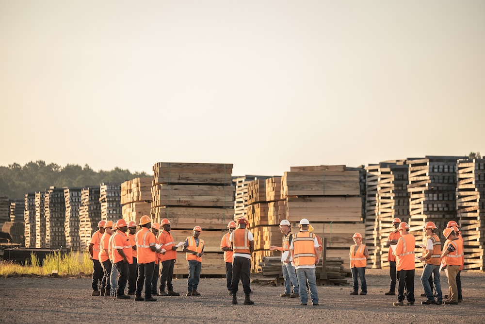 A crowd of workers in orange safety vests and hard hats stand meeting in a gravel lot in front of large stacks of wood.