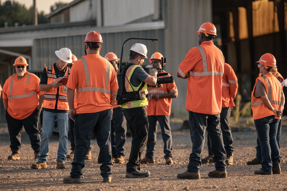 A crowd of workers in orange safety shirts and hats stand around a gravel lot in a meeting while a camera man films from the middle of the group.