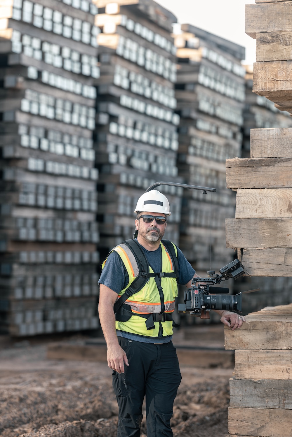 A camera operator with sunglasses stands amid pallets of wood in an industrial setting.