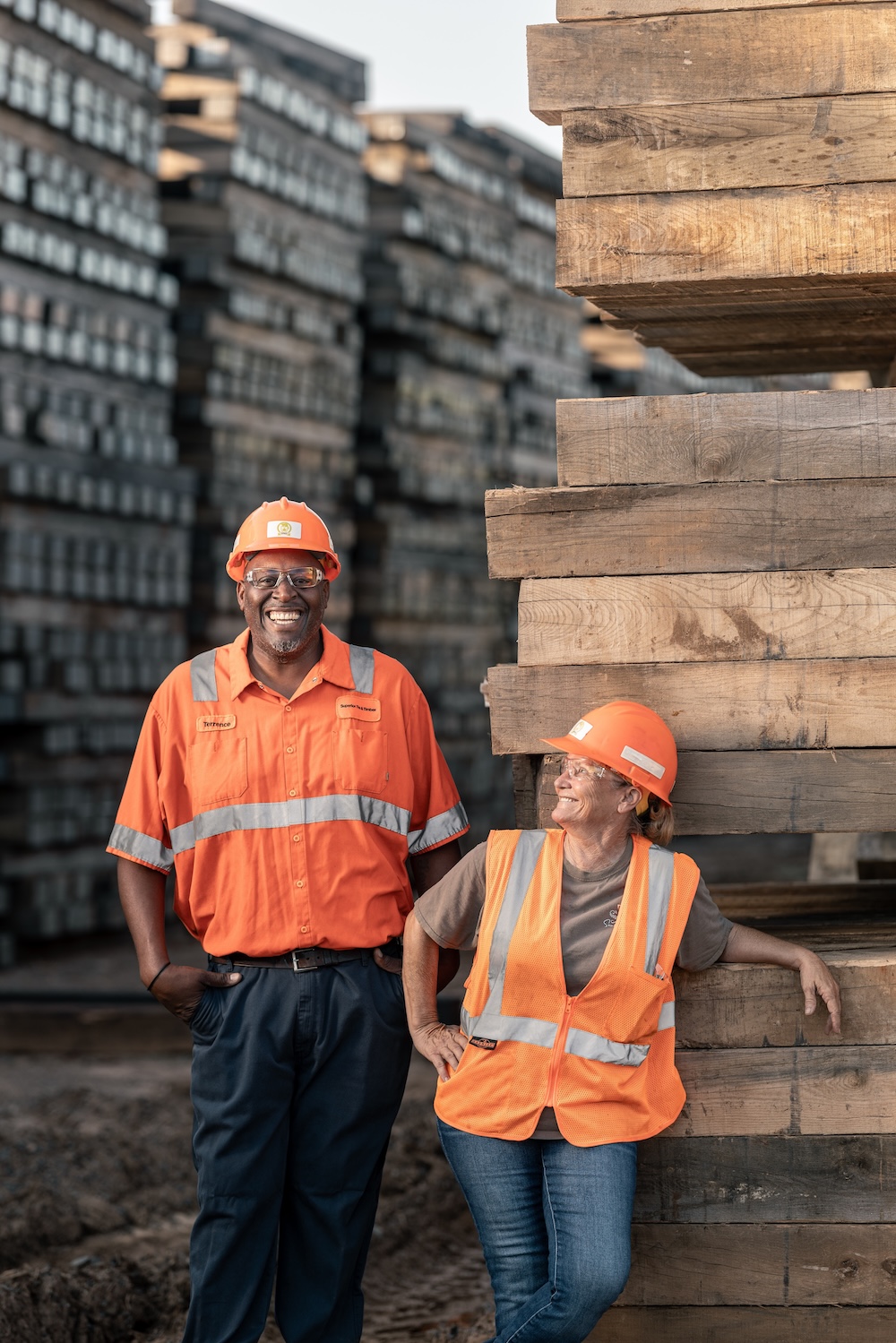 Two workers in safety gear smile and laugh while standing in front of tall stacks of timber.