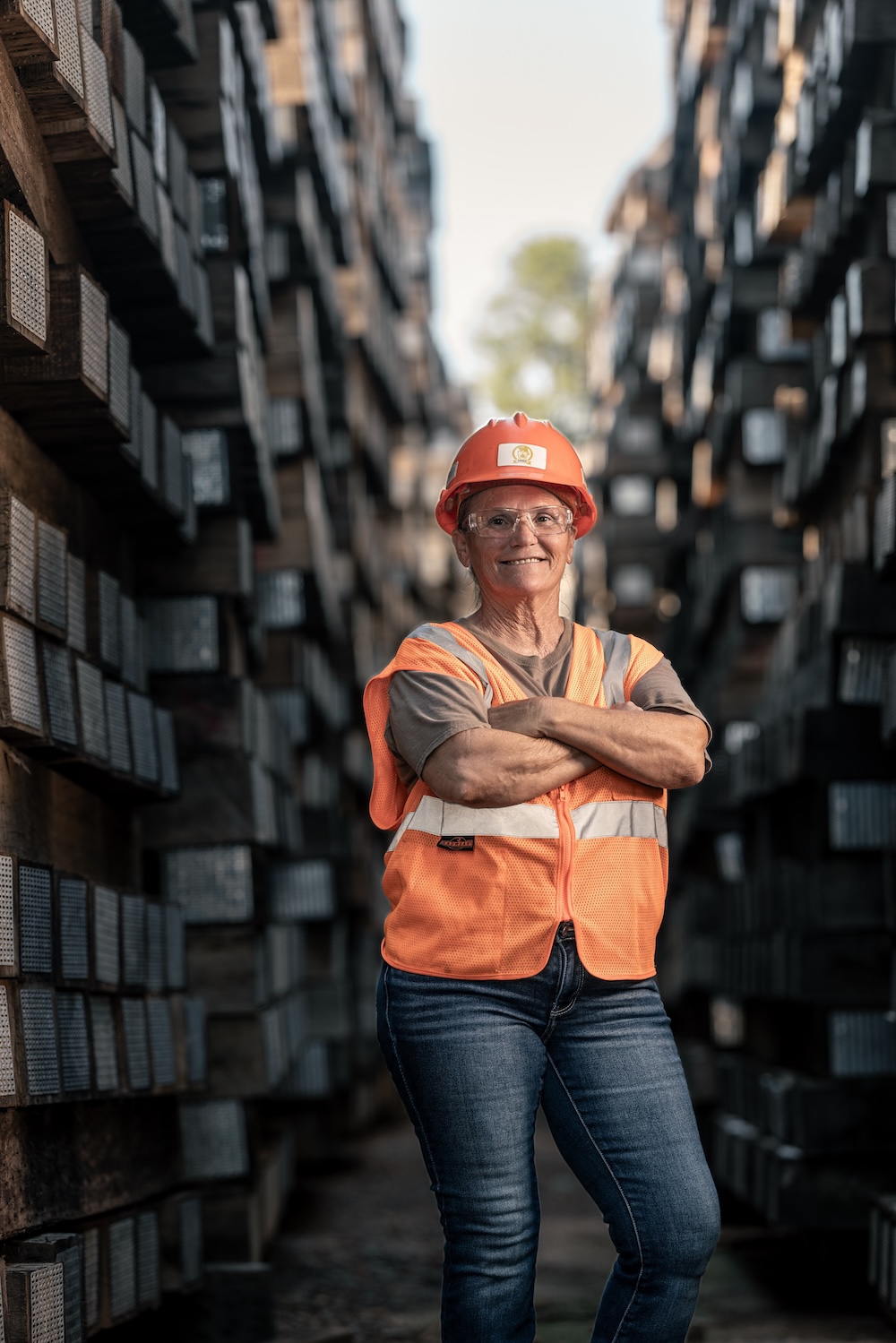 A female worker in an orange safety vest, hard hat, and safety glasses smiles and folds her arms across her chest as she stands among tall stacks of timber.