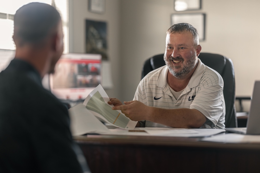 A businessman behind a desk smiles at another businessman and points at some paperwork.