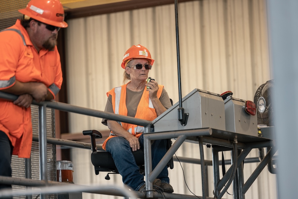A woman in an orange safety vest and hat holds something in her hand and sits behind industrial machinery controls while another worker looks on.
