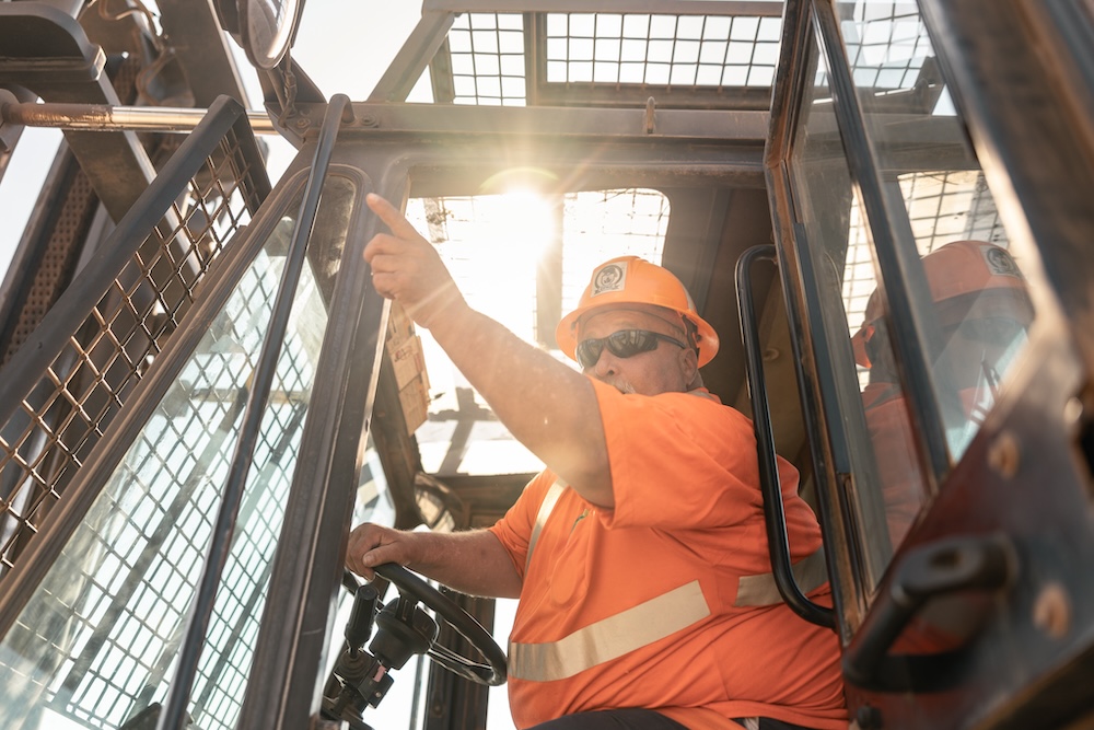 A man in an orange safety shirt and hat sits in the driver's seat of some type of vehicle, pointing out into the distance.