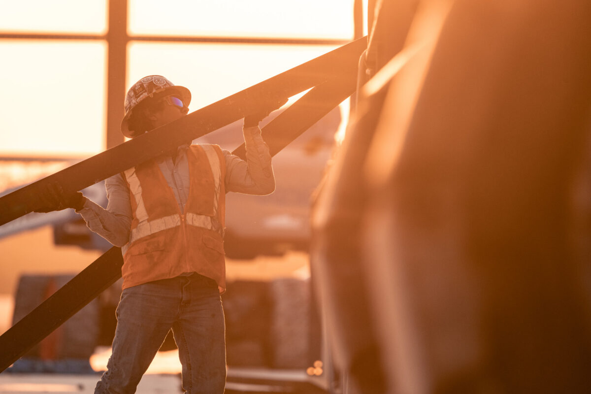 Golden hour shot of an employee in safety gear lifting a large piece of timber onto an out-of-focus piece of equipment.
