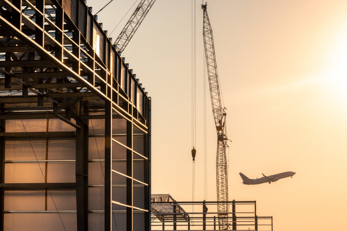 Backlit sunset image of a large open-walled warehouse with a large crane in the distance and an airplane in the background