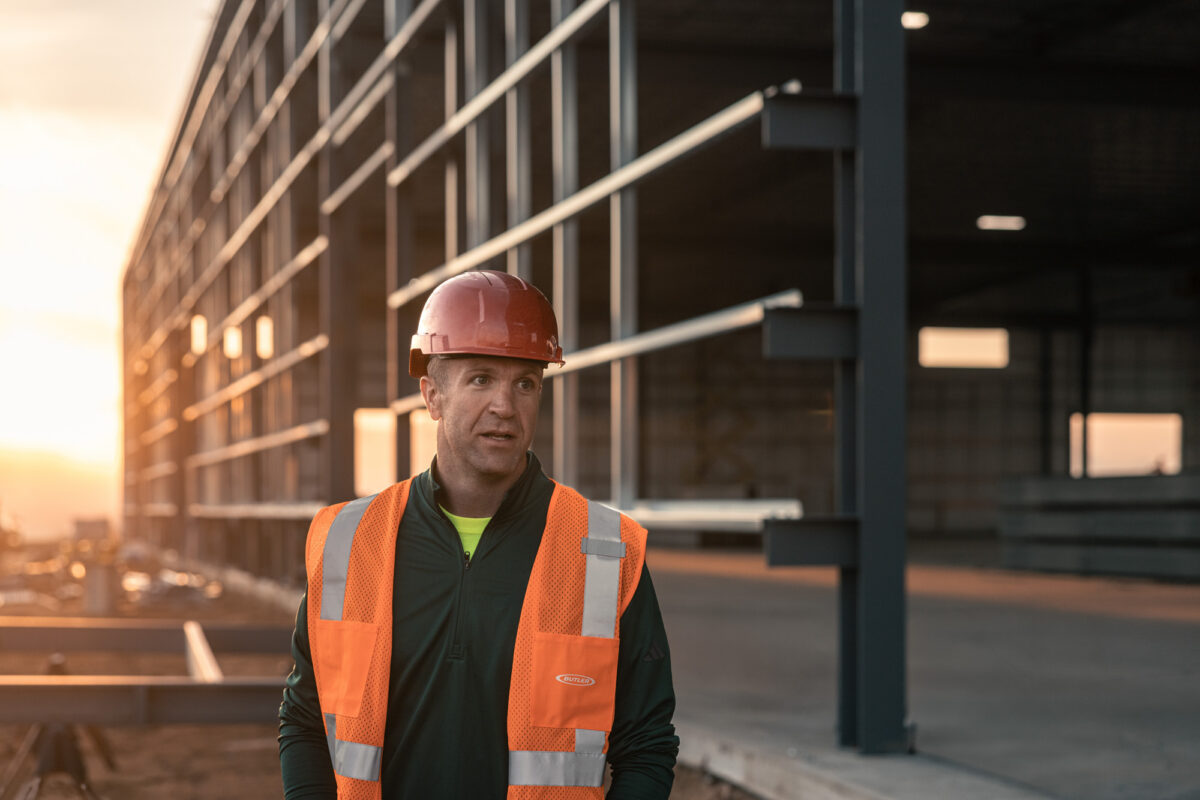 A man in a safety vest and red hard hat stands in front of a warehouse under construction.