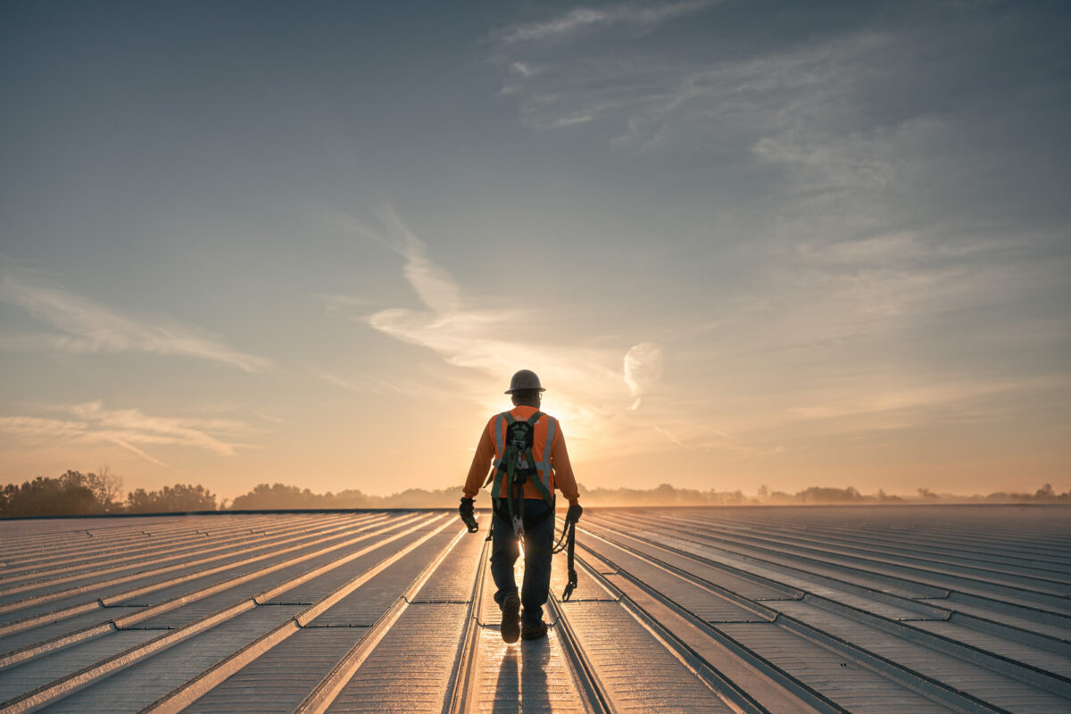 A man in safety gear walks away from camera atop a metal roof, as the sun sets over the tree line in the distant background.